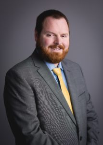 Formal picture of man with short hair and beard, dressed in gray suit with blue shirt and yellow tie.