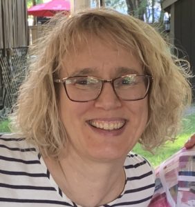 Close up photo of woman with glasses, blond curly shoulder length hair.  She is wearing a white and navy striped shirt.  